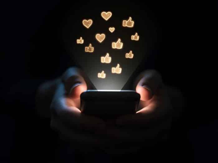 hands holding a smartphone in darkness, with glowing icons of hearts and thumbs-up floating from the screen, conveying social media engagement.