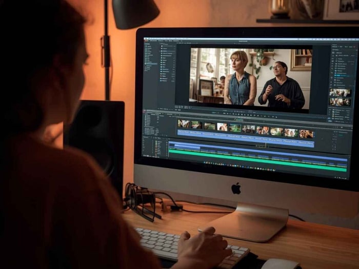 a person is seen editing a video on an imac, displaying editing software with timelines and a scene of two people talking. the room has a warm, focused atmosphere.
